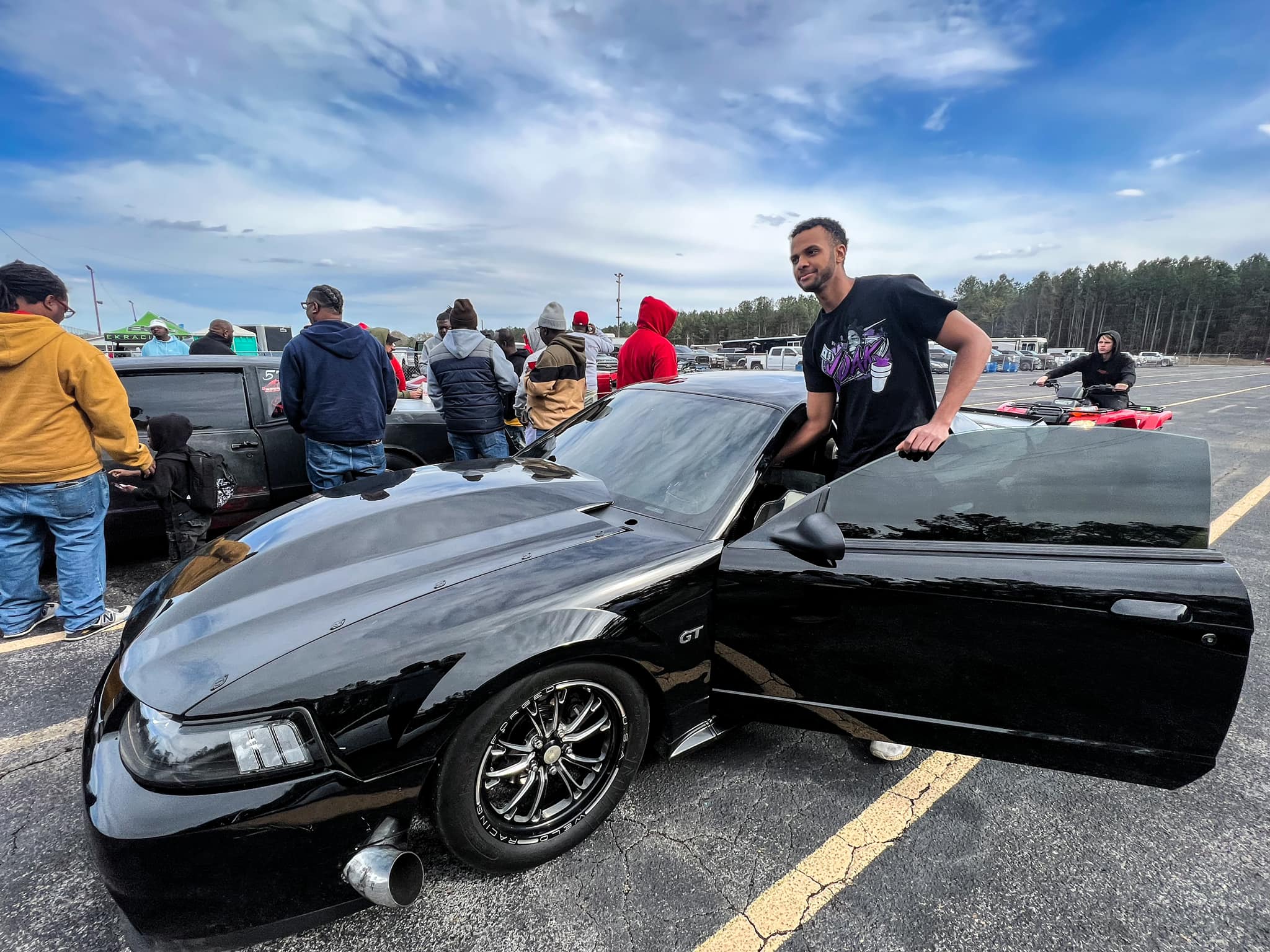 Black car with open door in a parking lot with people around